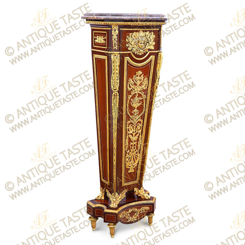 Louis XVI style ormolu-mounted pedestal after the model by Jean-Henri Riesener, late 19th century, with stepped veined marble top & canted front corners above an Egg-and-Dart ormolu trim, on a rectangular concave upper section followed and centered by a Mercury sunburst ormolu mask with crossed lower branches, the angles with an acanthus-cast scrolled volute hung and foliate festoons, above the tapering upright, the paneled front finely chiseled and burnished with a flower-filled Rinceau style vase ormolu pattern flanked by scrolling foliage and floral swags issuing suspending festoons above a Lyre and crossing blowing instruments with surrounding wreath, all placed on a pomelle bubinga veneer background within an ormolu beaded trim, on winged lion-paw feet and shaped plinth on toupie leafy feet
